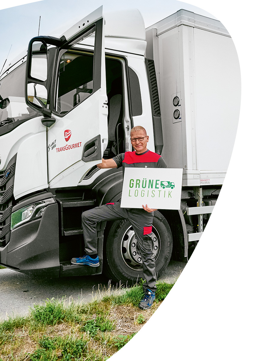 Transgourmet Fahrer steht neben dem Führerhaus seines LKW und hält ein "Grüne Logistik"-Schild in der Hand