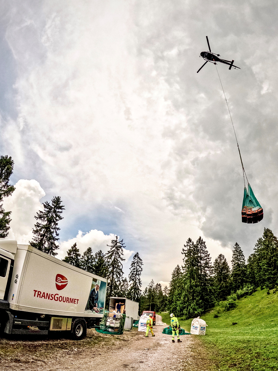 Ein Transgourmet-LKW entlädt an einem Hang Ware. Ein Helikopter transportiert diese einen Berg hoch.